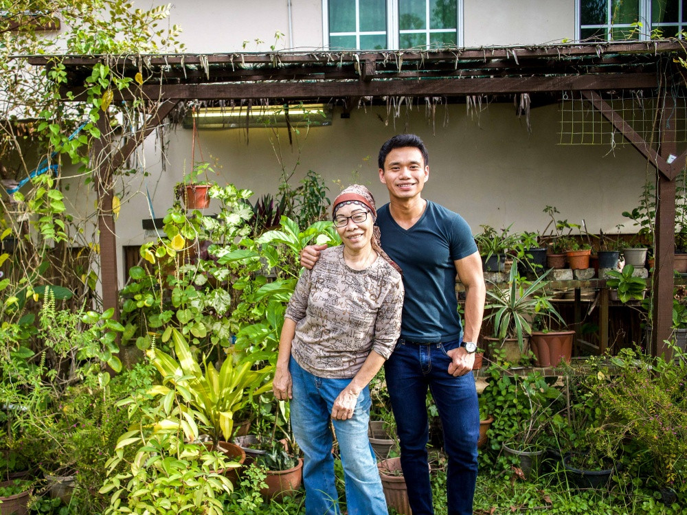 Pn Norhanida dan Hazli berdiri di hadapan rumah mereka dengan taman penuh sayuran dan buah-buahan. Kebanyakan daripada buah, sayuran dan ulam yang ditanam di sini dipetik untuk The Sixpack Baker. Gambar ihsan: Natasha Sim